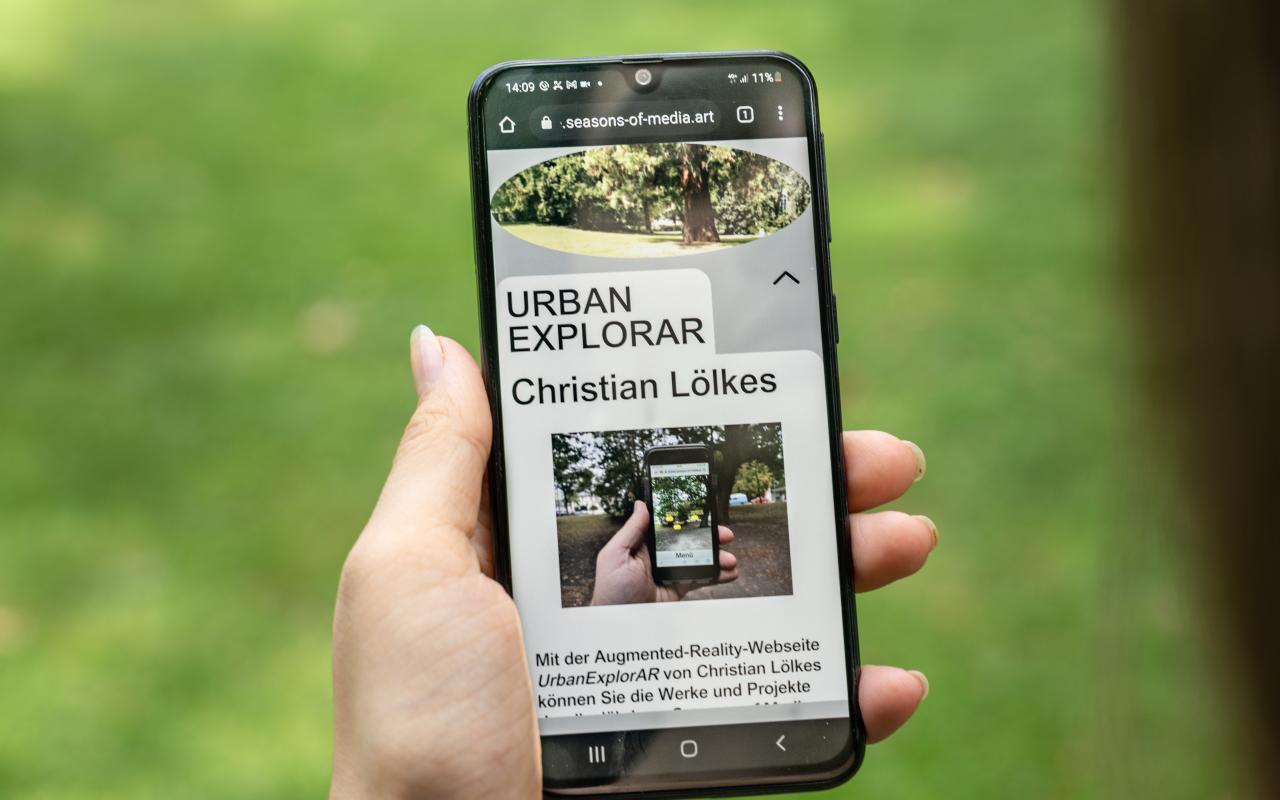 The picture shows a hand holding a smartphone with the work »UrbanExplorAR« on it.