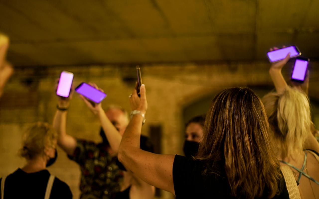 Zu sehen sind mehrere Personen in einem abgedunkelten Raum, die ihre Arme nach oben strecken. In ihrer Hand halten sie ein Smartphone, dass pink aufleuchtet.