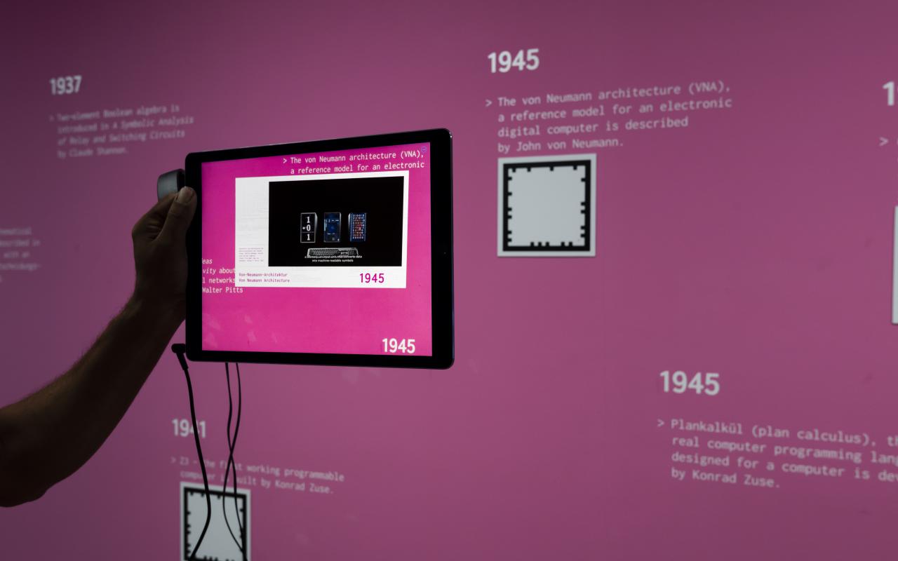 A hand holds a tablet in front of a pink wall. 