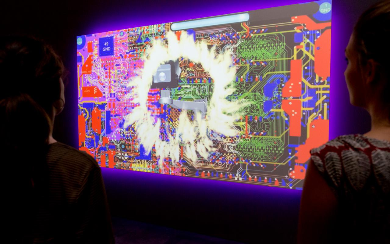Zwei Besucherinnen betrachten ein Video-Kunstwerk, das eine Collage aus Computer-Hardware und Flammen zeigt