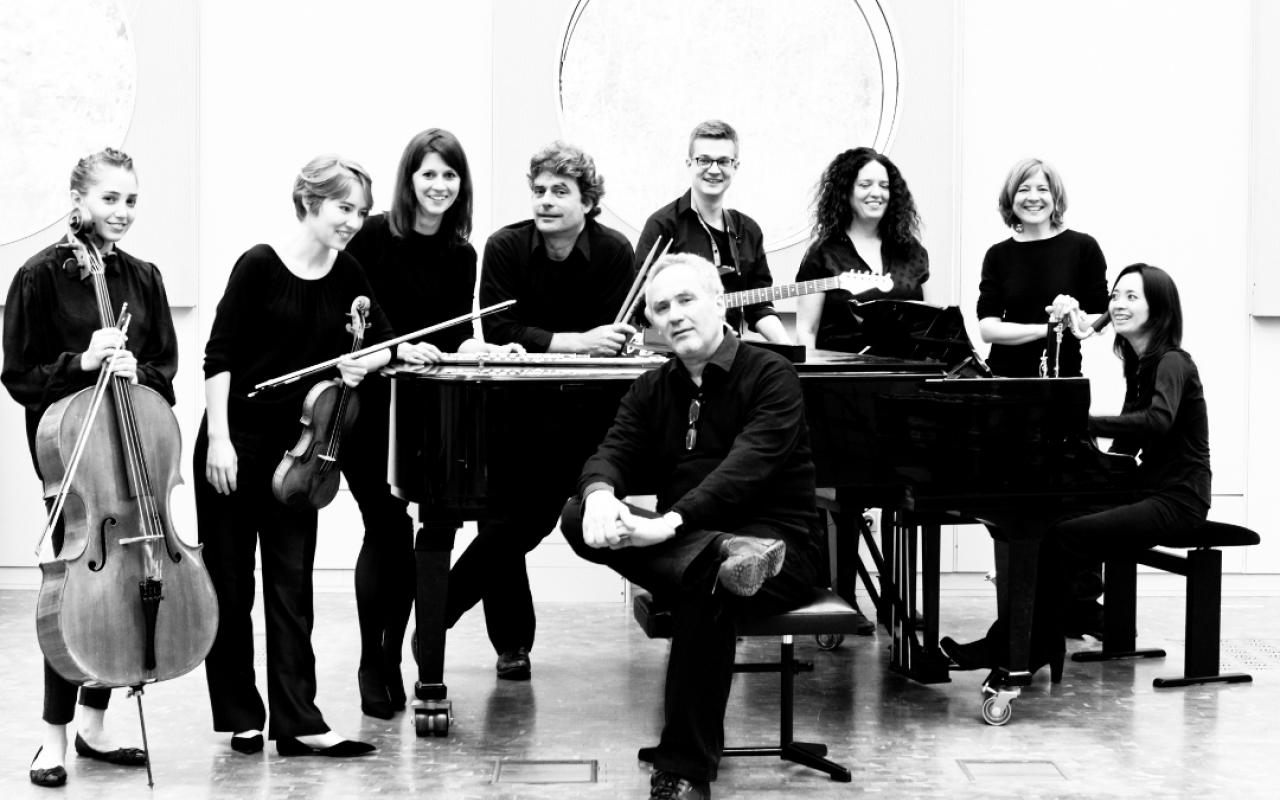 On display is the "Ensemble Experimental" consisting of eight people. One person sits in front of a piano and the rest stand behind it. The picture is black and white.