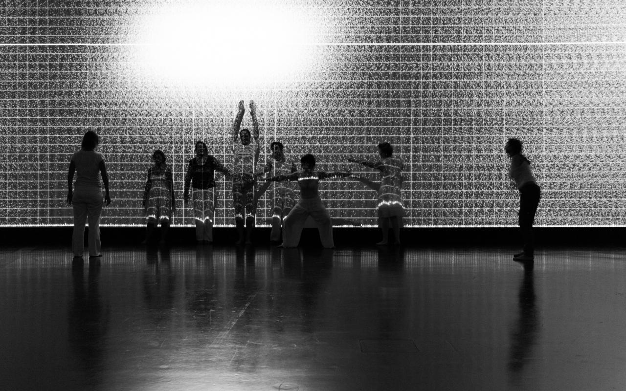 A group of dancers is moving in front of a wall with black and white projections.