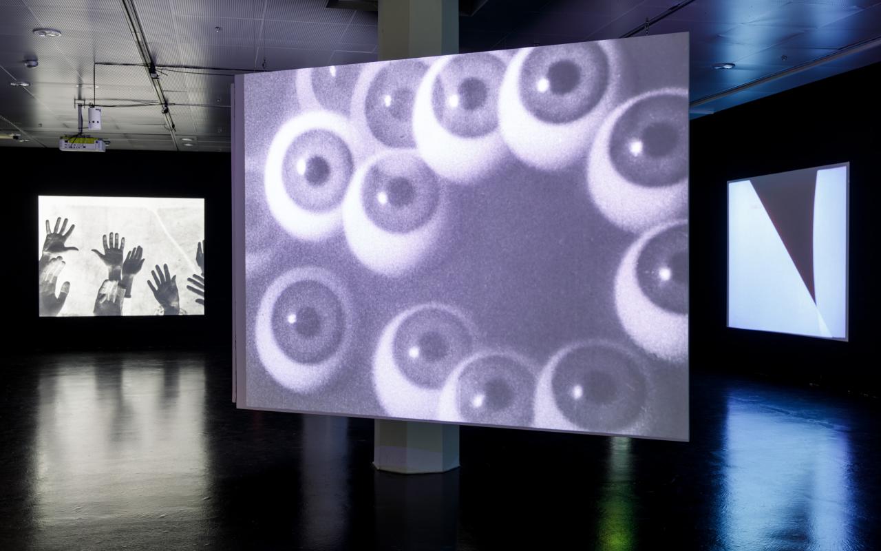 The photo shows the exhibition room with a large canvas in the middle, depicting spheres that look like eyeballs. To the left and right there are two other canvases in the background, on which hands and black and white shapes are depicted. 