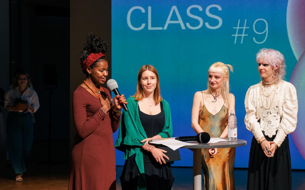 Four people on stage at the [MASTERCLASS] #9 opening. The person on the far left is holding a microphone in one hand and has the other hand on her heart.
