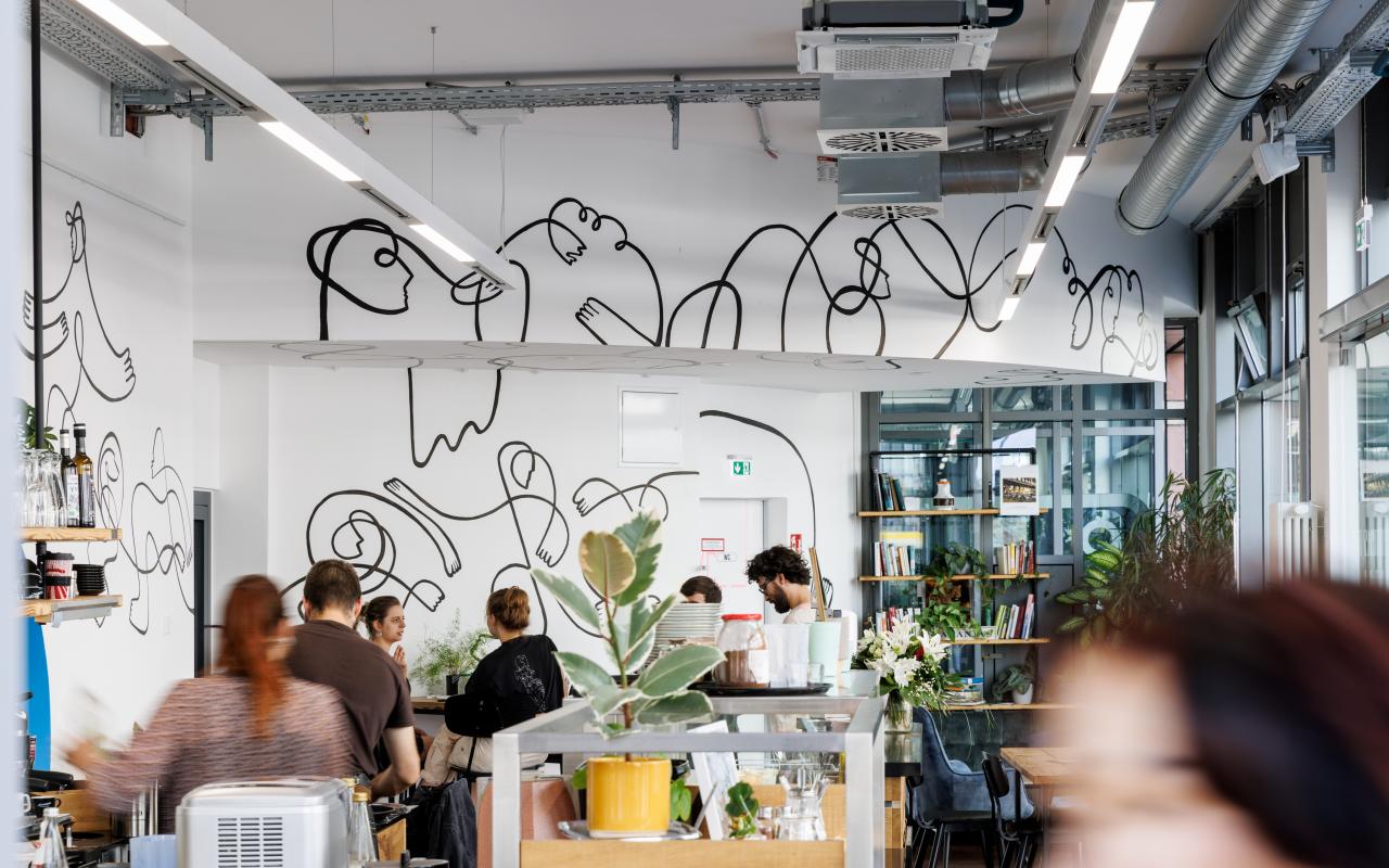 Auf dem Bild ist das Kunstwerk von Alina Bukina »One Line Series«, im intro Café am Kronenplatz zu sehen. Eine schwarze Linie zieht sich an den Wänden durch das ganze Cafe. Im Café selbst sitzen einige Menschen.