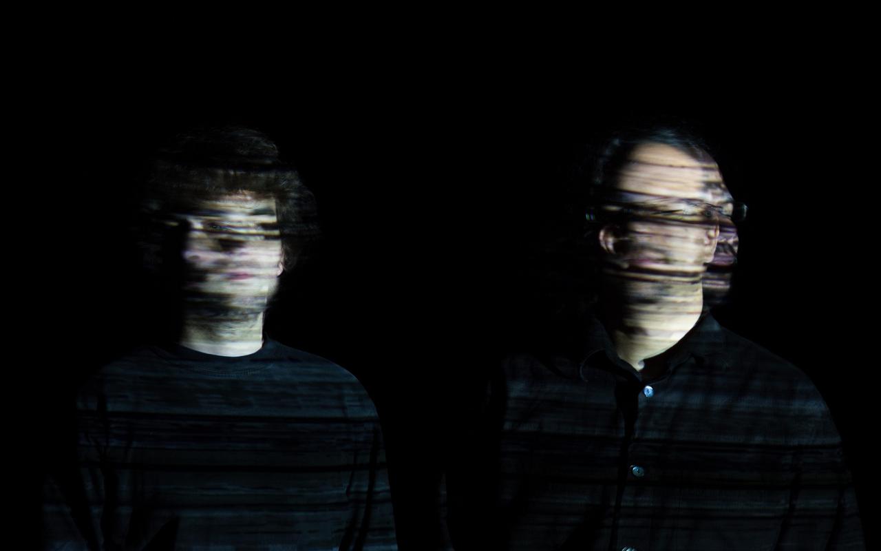 You can see two men in a very dark room. Only their faces can be recognized, although even these are only blurred. This effect is created by moving the head from left to right during a long exposure time.