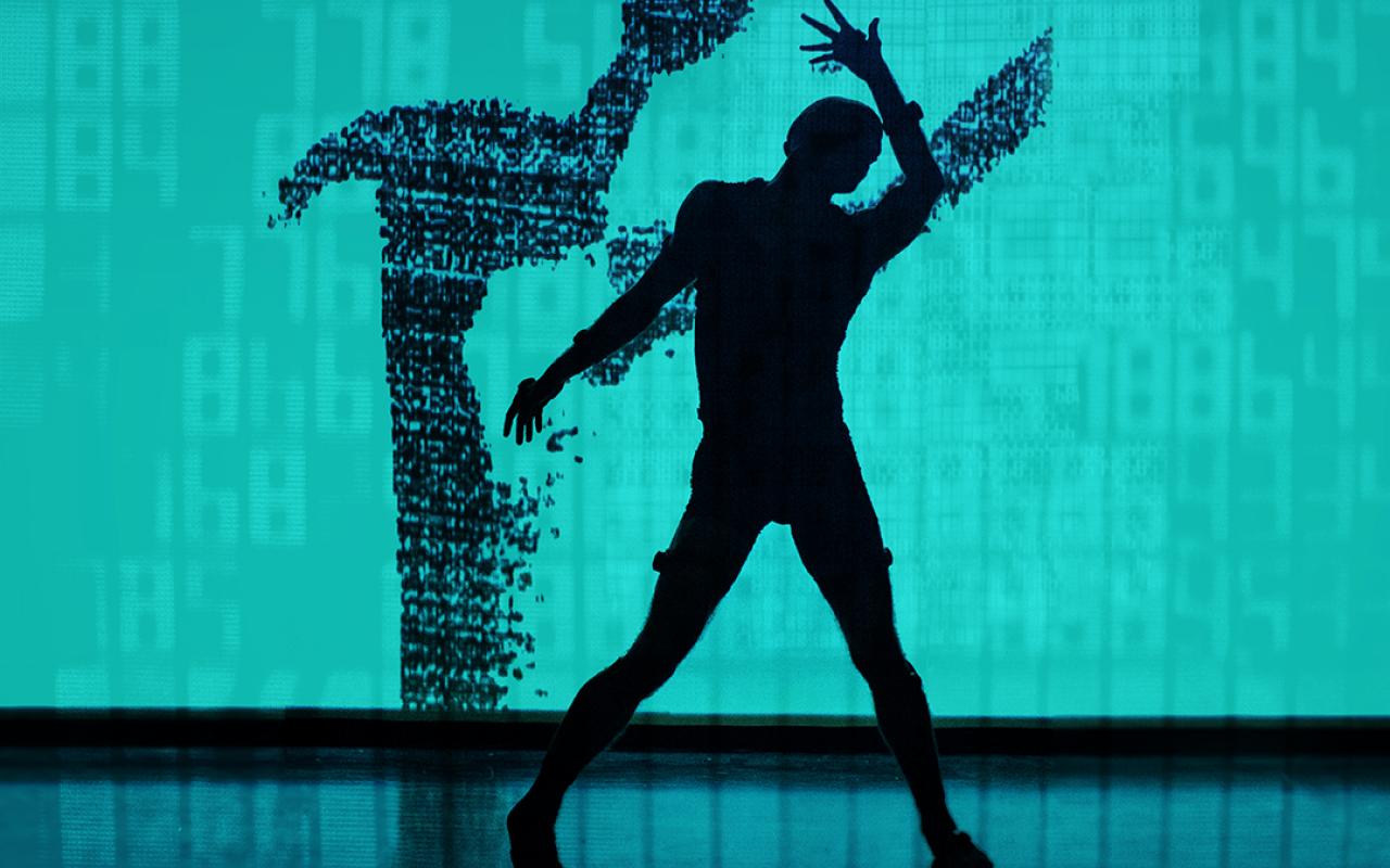 The photo shows a stage with turquoise background and black floor, on which the silhouette of a dancing man can be seen, whose shadow is depicted on the back wall.