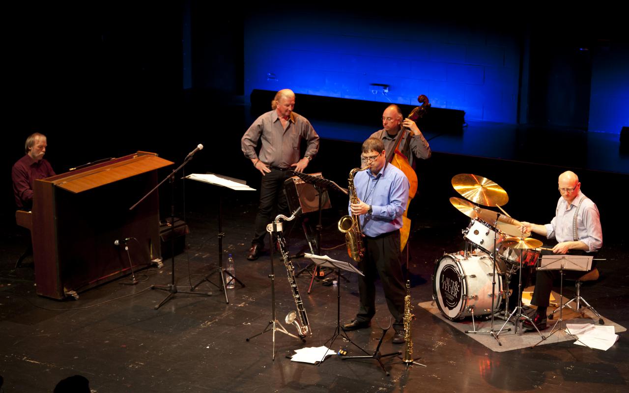 Five musicians at a stage situation can be seen.
