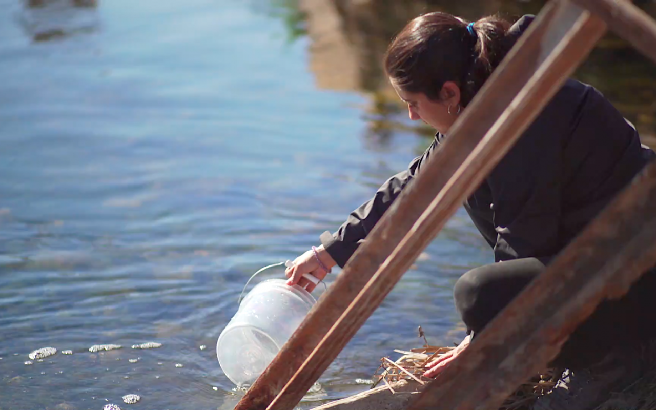 Einzelbild eines der beiden Videos des Werkes Hidroscopia Loa. Man sieht wie die Künstlerin Claudia González Godoy eine Wasserprobe aus dem Fluss Loa entnimmt.