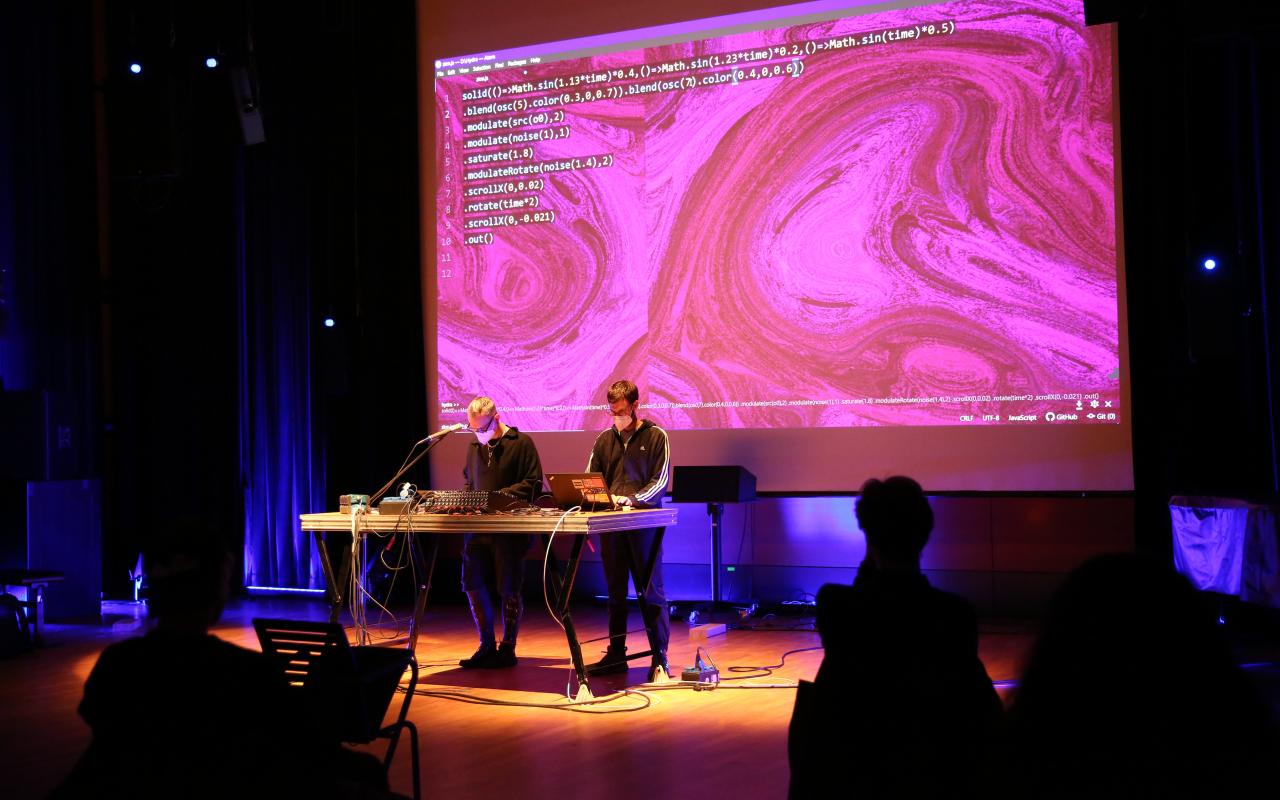 The photo shows two men standing in front of a large screen with a desk and laptop in front of them. On the canvas you can see a large pink background and white writing. A few people from the audience can be seen from behind.