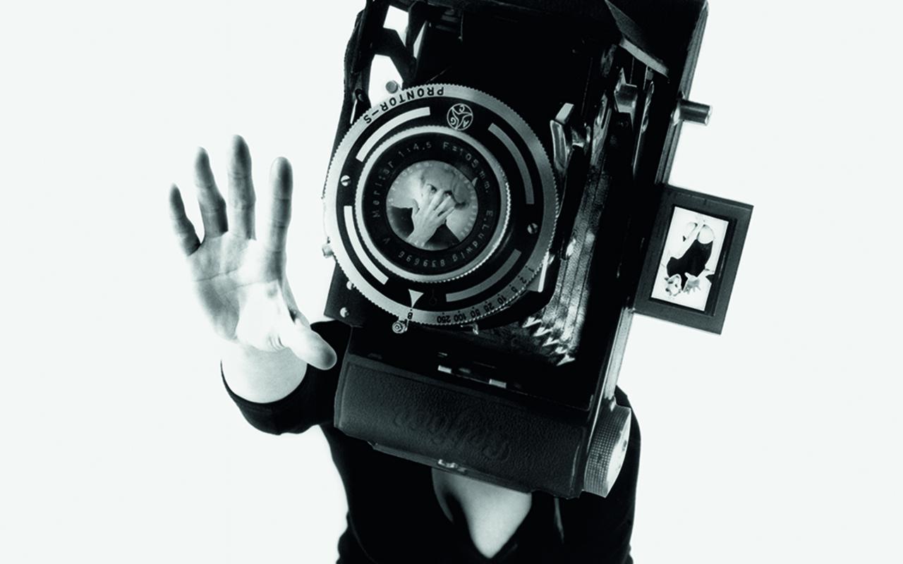 Das Schwarzweiß-Photo zeigt eine Frau in einem schwarzen Minikleid auf den Knien. Ihr Gesicht ist von einer überdimensional großen Kamera verdeckt. 