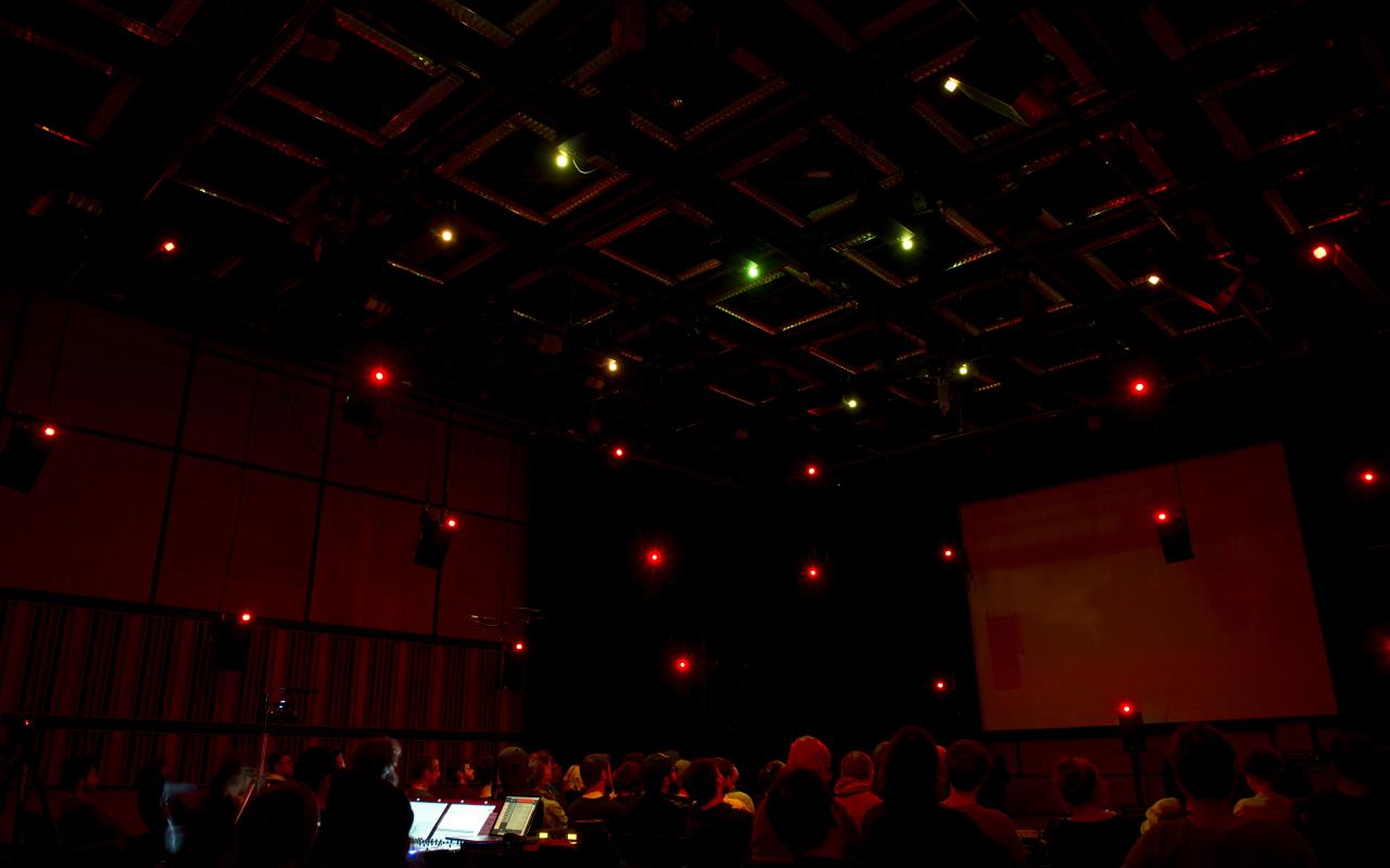 A dark large room with many sitting people. There are single lights on the ceiling.