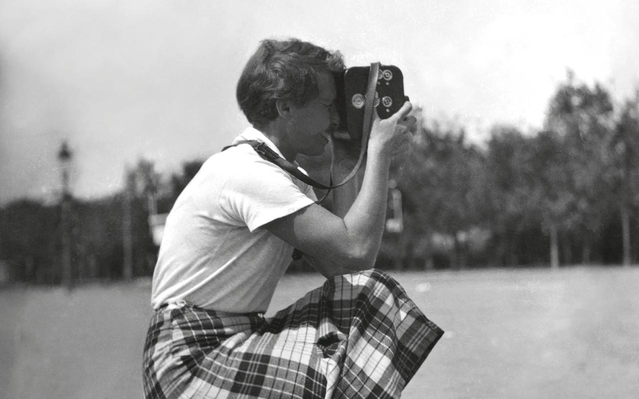 Die Schwarz-Weiß-Fotografie zeigt eine Frau in der Hocke mit einer Kamera.