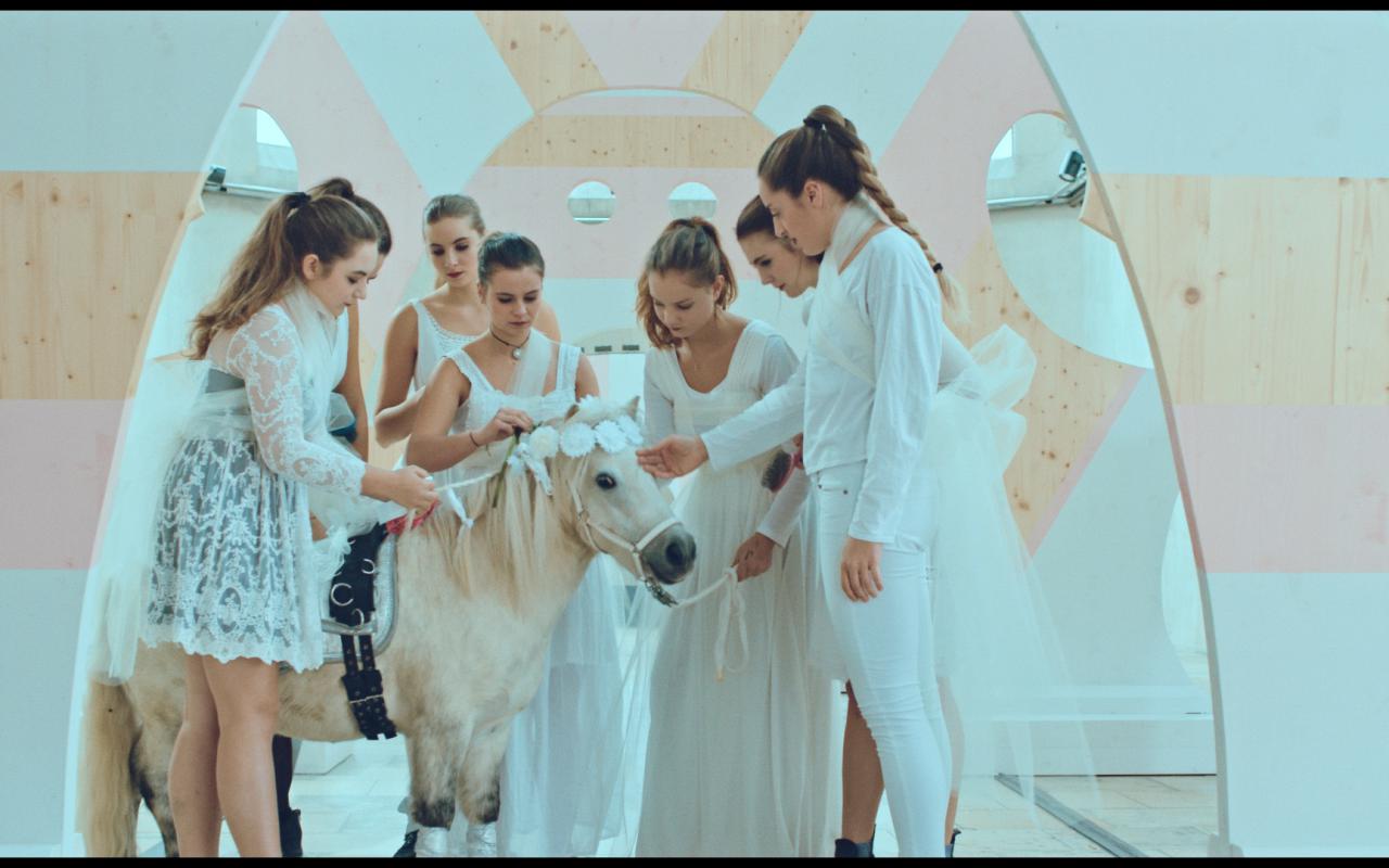 The photo shows a white pony surrounded by seven girls in white clothes.