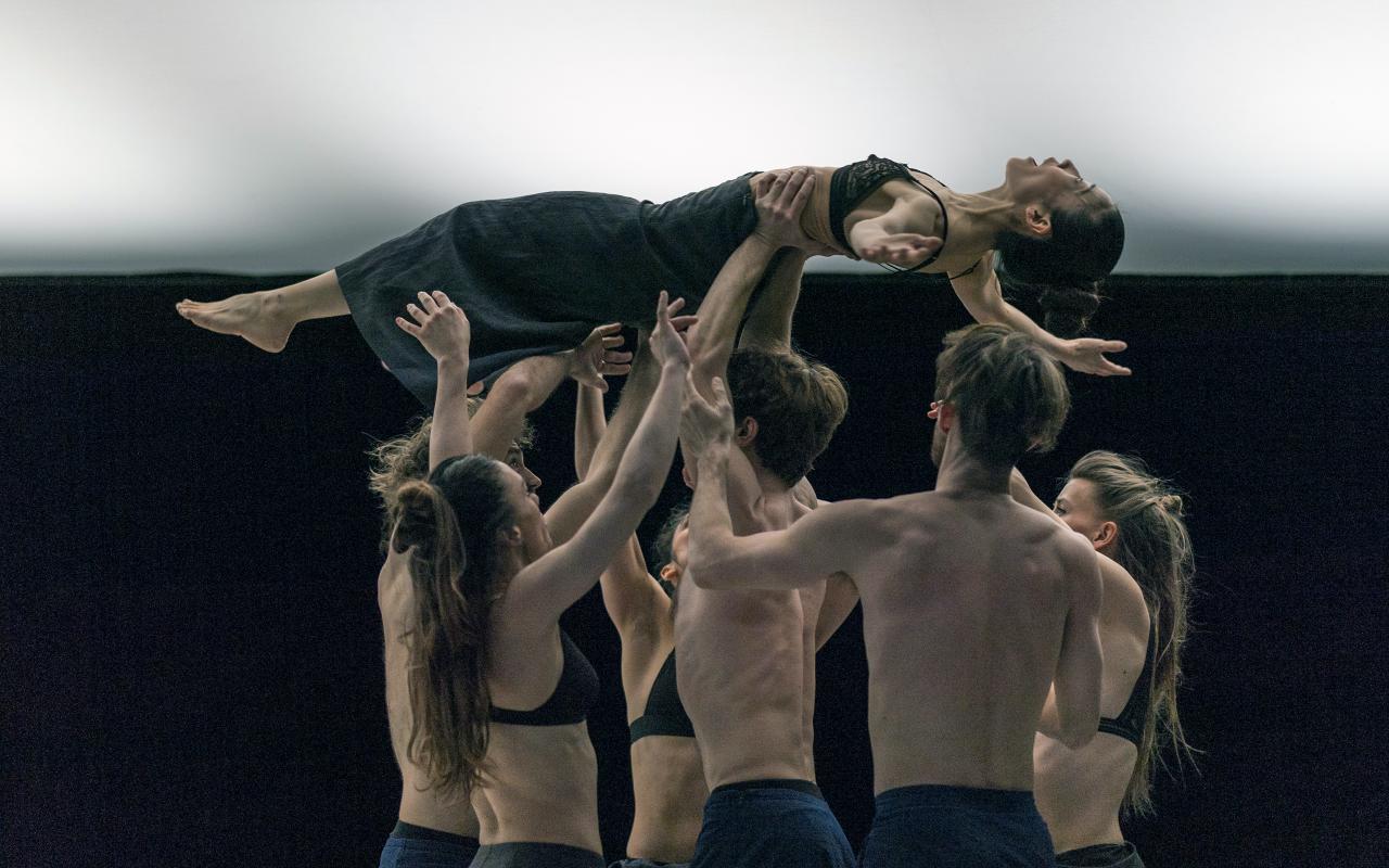 Six dancers hold a dancer above their heads. The dancers are topless and wear dark trousers.