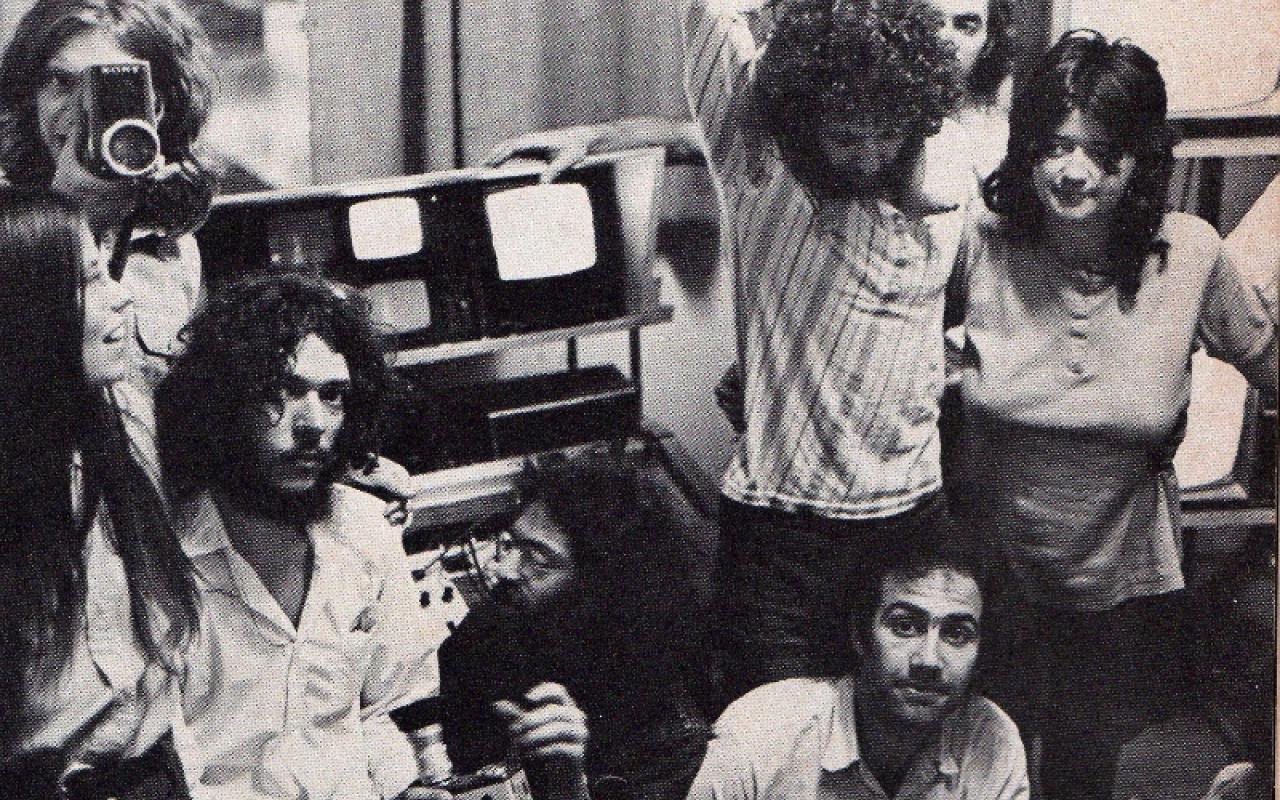 The black-and-white image shows a group of long-haired young people in front of small screens