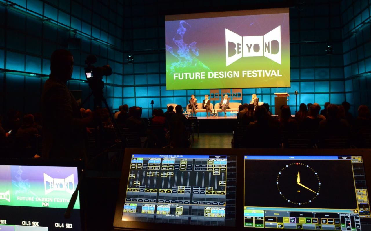 View into a large darkened room. On the opposite wall is a large screen with the logo of the Beyond Future Design Festival.