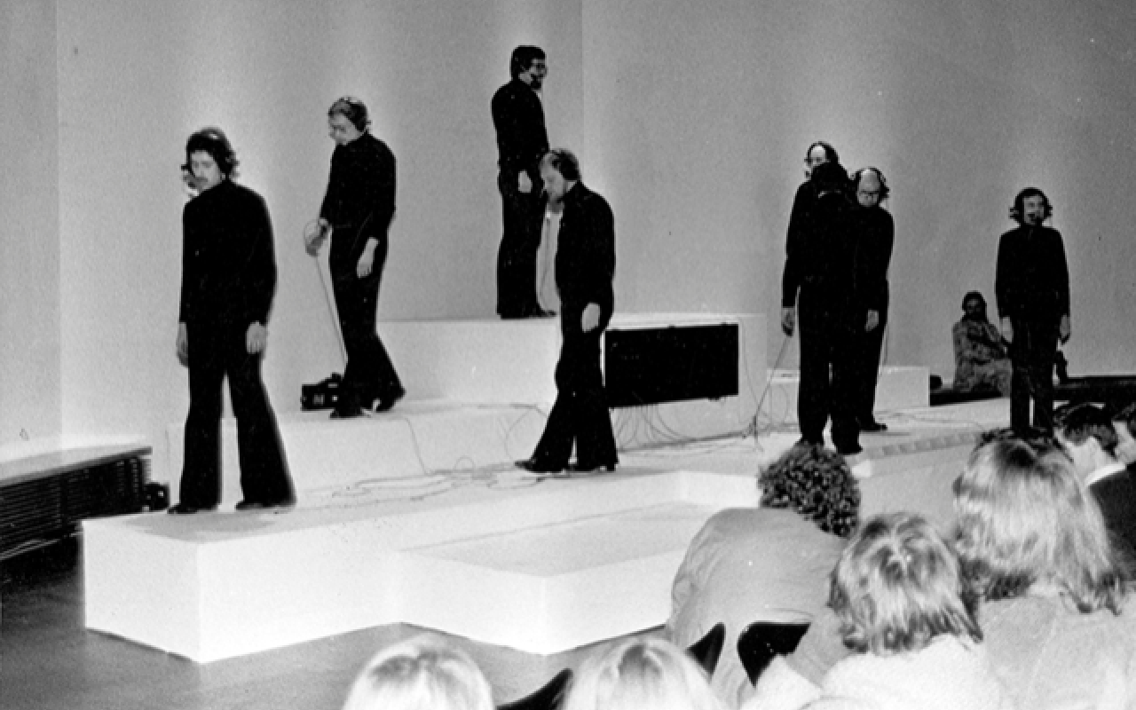 Es sind neun in schwarz bekleidete Personen zu sehen, die auf Podesten stehen. 