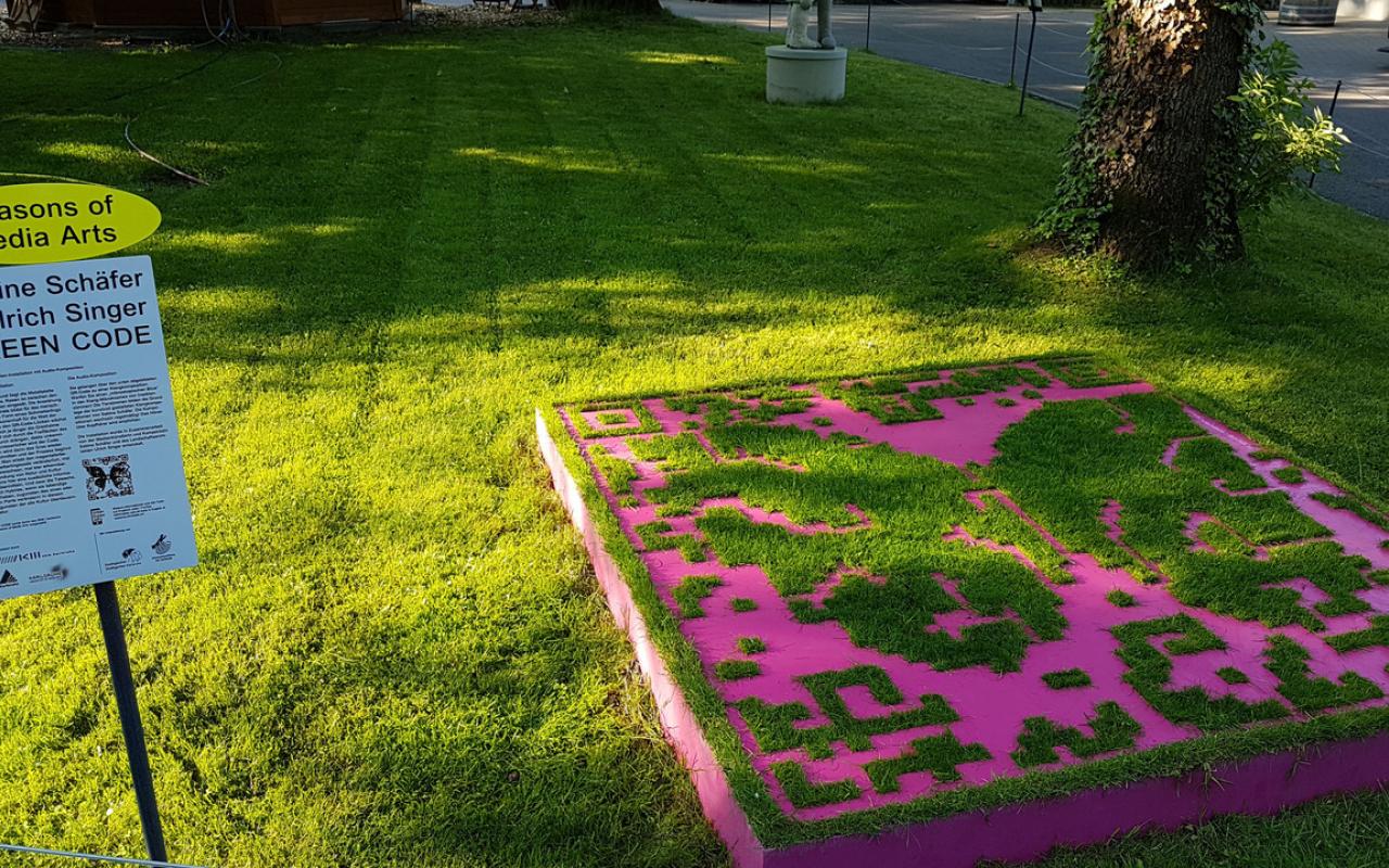 You can see a stencil lying on a surface of grass. The template has the form of a QR code. Grass grows through the free areas of the stencil.
