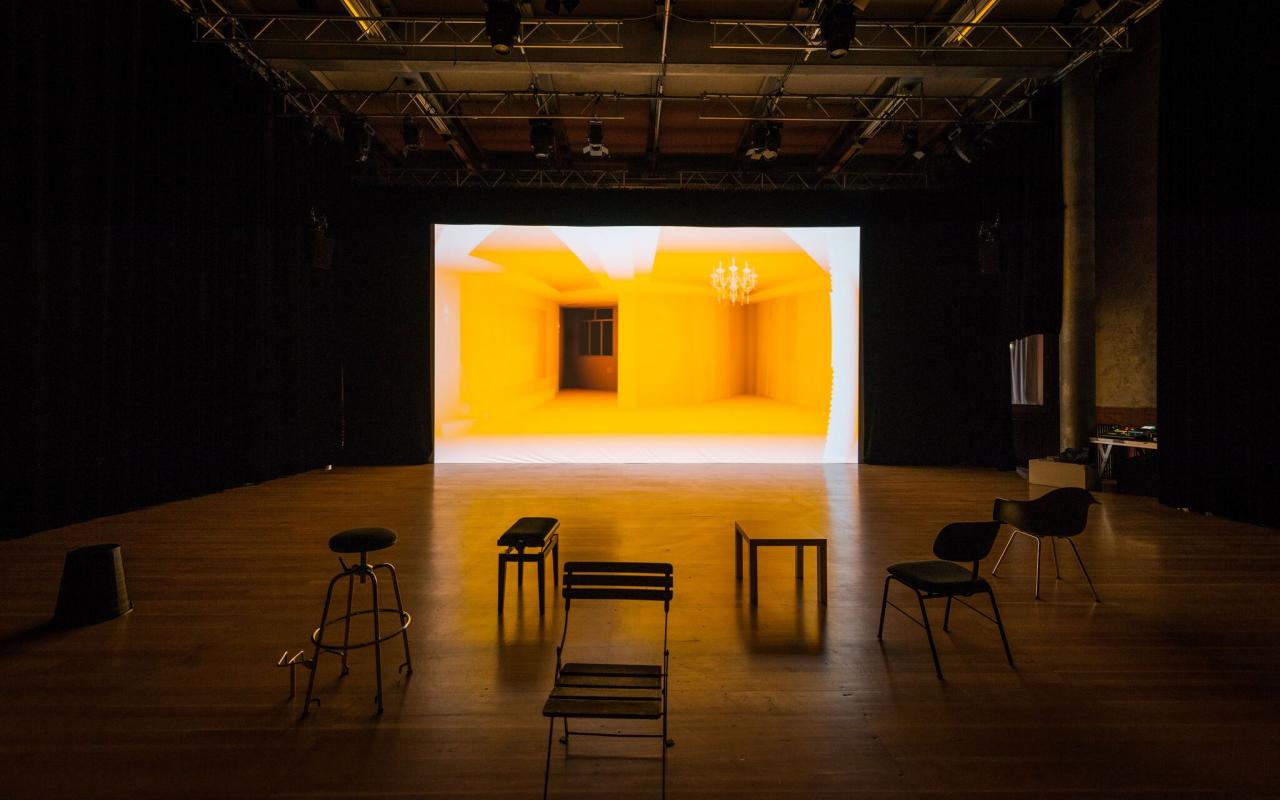 Ausstellungsansicht des Werks »Homeschool« von Simone C. Niquille / Technoflesh in einem abgedunkelten Raum, einigen Sitzgelegenheiten und einer großen Leinwand auf die ein gelber Innenraum projiziert wird