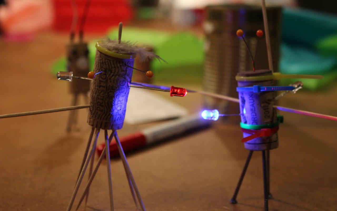 Zwei Korken, die zu kleinen Figuren umgewandelt wurden und mit LEDs ausgestattet sind stehen auf einem Tisch mit allerlei Bastelutensilien.
