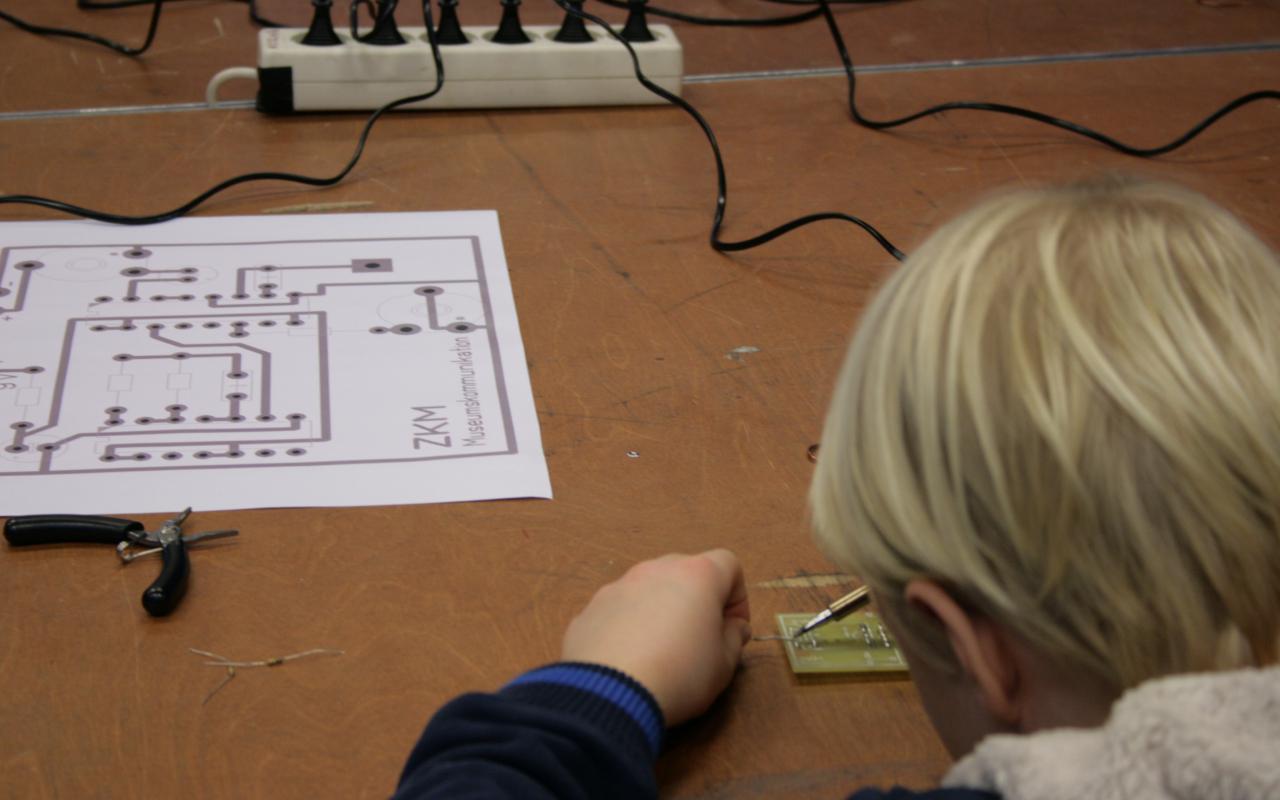 A boy is working on a circuit board with a soldering iron, out of which he is buil
