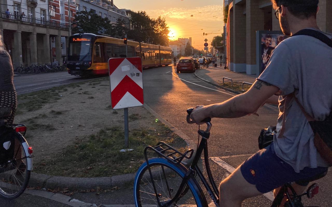 In the foreground, a young man on a bicycle is riding along a road. In front of him, the setting sun is shining and illuminating the streetcar that crosses the landscape.