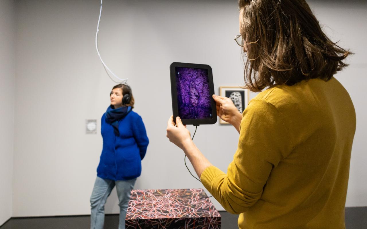 to see are two women. One is standing in the background and is photographed by the one in front through a tablet.