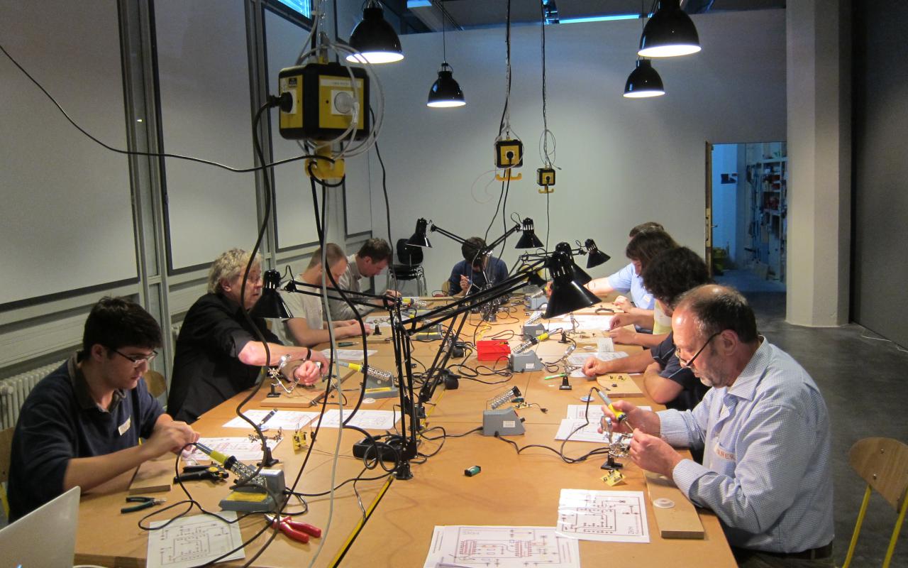 An einem riesigen Tisch sitzen viele Workshopteilnehmer, die mit Lötkolben arbeiten und es liegt allerlei Material kreuz und quer herum.