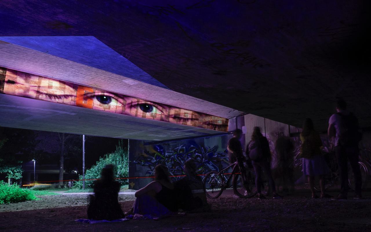 A bridge in a park at night. A row of pairs of eyes is projected in a horizontal strip on the outer wall of the bridge. The rets of the bridge shine in different colors.