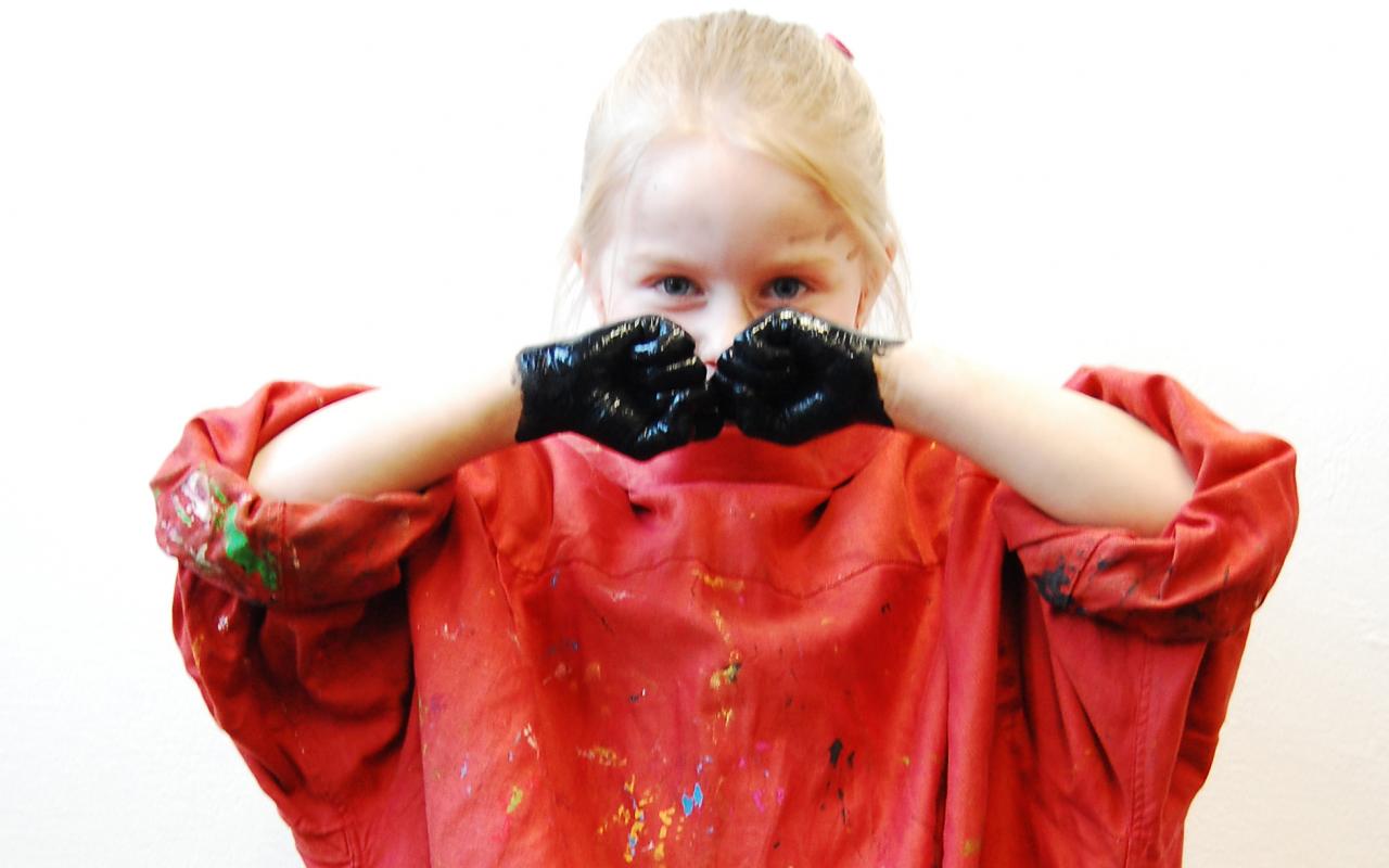 Ein junges Mädchen schaut direkt in die Kamera, dabei hält es die schwarzen Fäuste vor das Gesichtes. Das Mädchen trägt ein leuchtend rotes Hemd, das mit Farbe bekleckert ist.