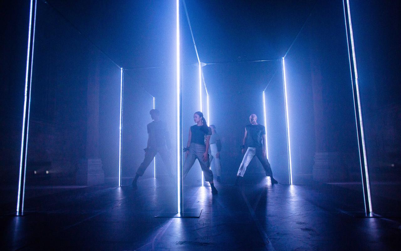 Eine Gruppe TänzerInnen steht in einem abgedunkelten Raum, der von blauen Neonröhren beleuchtet wird.
