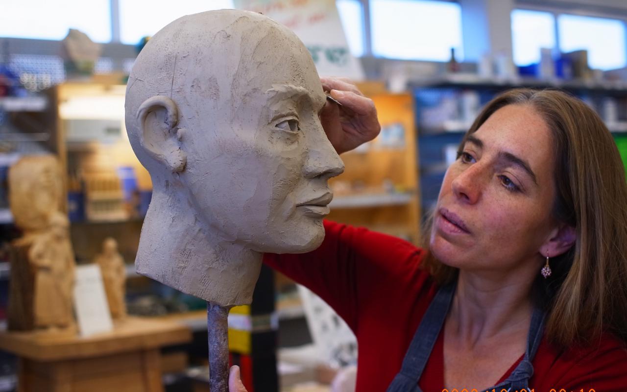 Ein Tonmodell eines Kopfes steckt auf einem Stab. Eine Frau arbeitet mit einem Modellierwerkzeug an der Augenpartie.