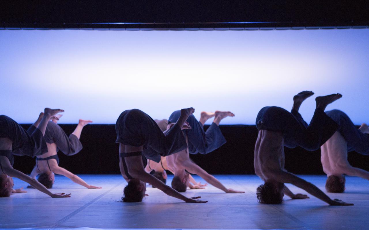 Seven different dancers have bent their legs in the headstand. In the background there is a large, milky screen. 