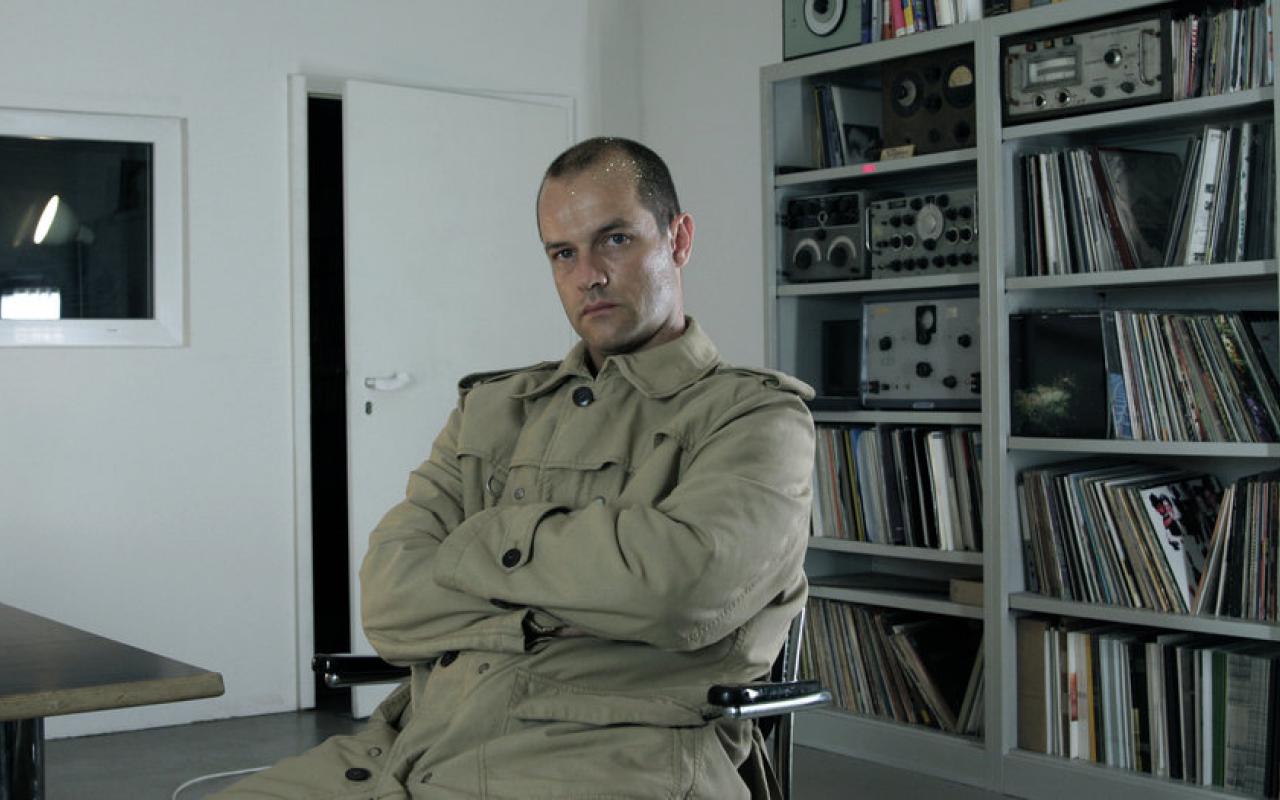 Der Wissenschaftler und Komponist Marcus Schmickler sitzt mit vor der Brust verschränkten Armen vor einem Regal mit Schallplatten und elektrischen Geräten