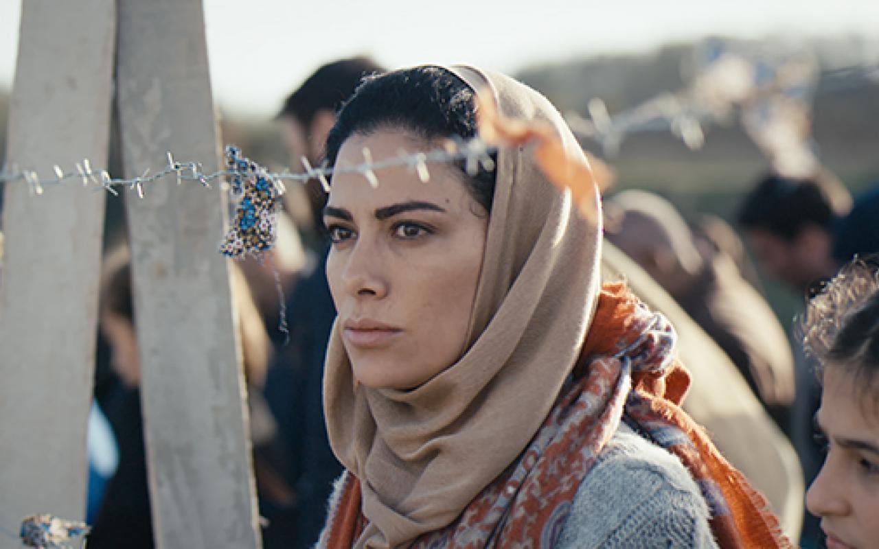 A woman with a headscarf is standing behind a barbed wire fence.