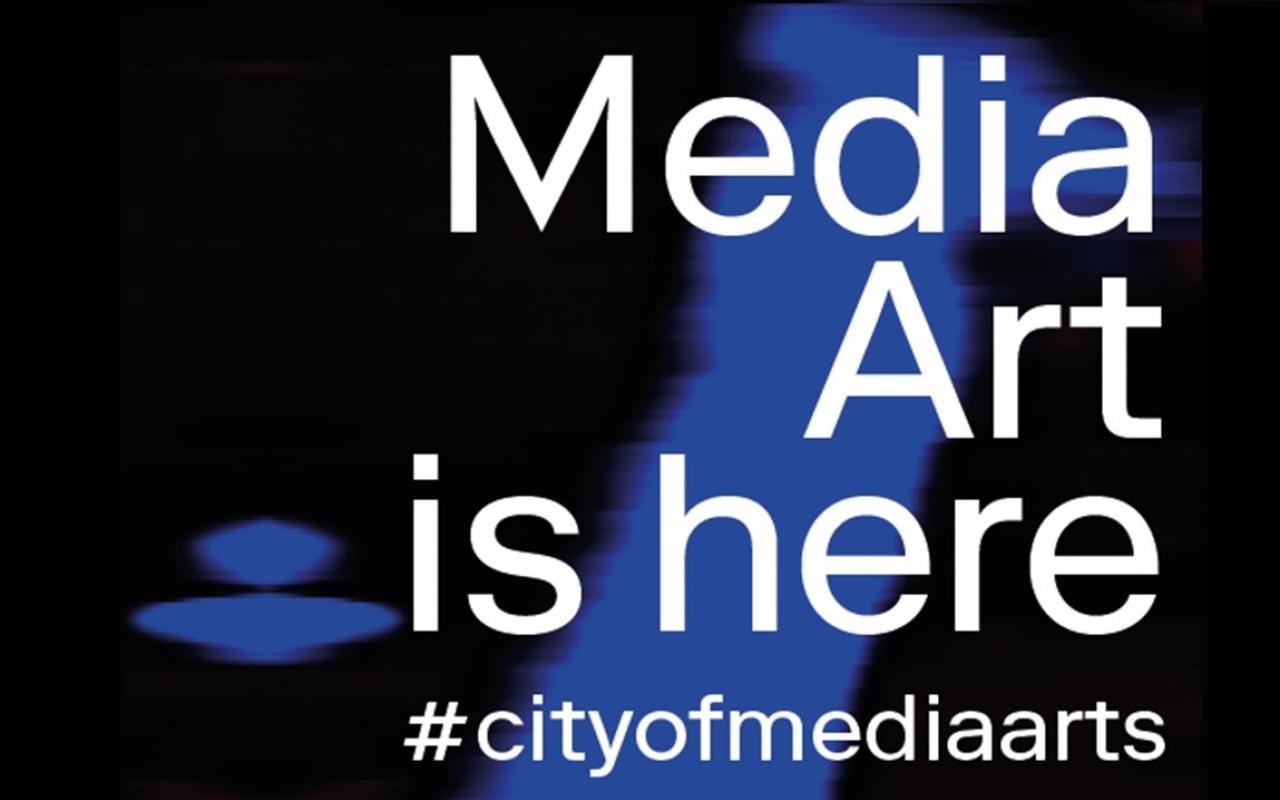 Zu sehen ist der Schriftzug Media Art is here #cityofmediaarts auf einem schwarzen Hintergrund, der von einem blauen Streifen durchzogen ist.