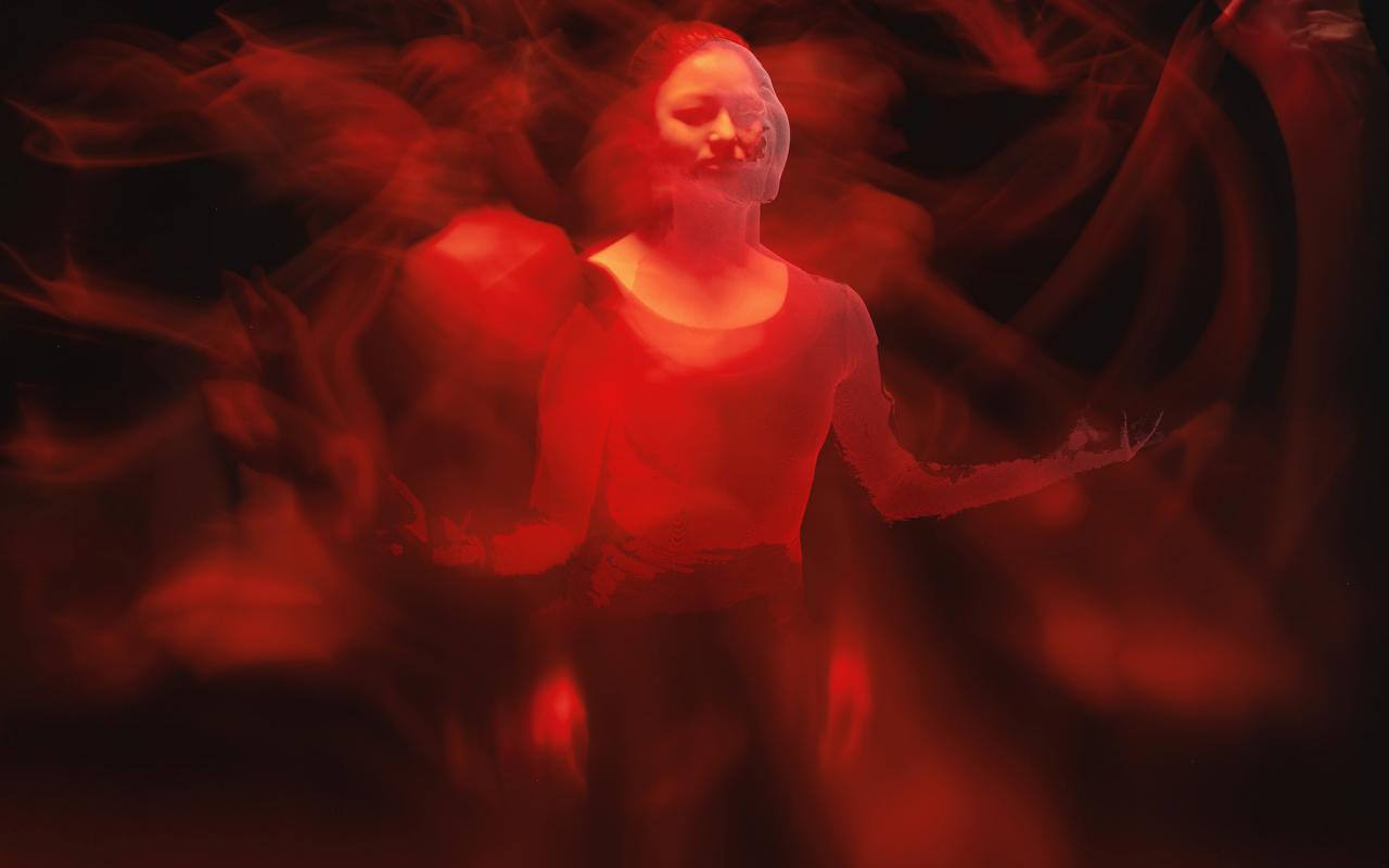 The photo shows a woman illuminated by red light in a darkened room. This expression shows several blurred arms and hands in motion, almost resembling an Indian deity. 