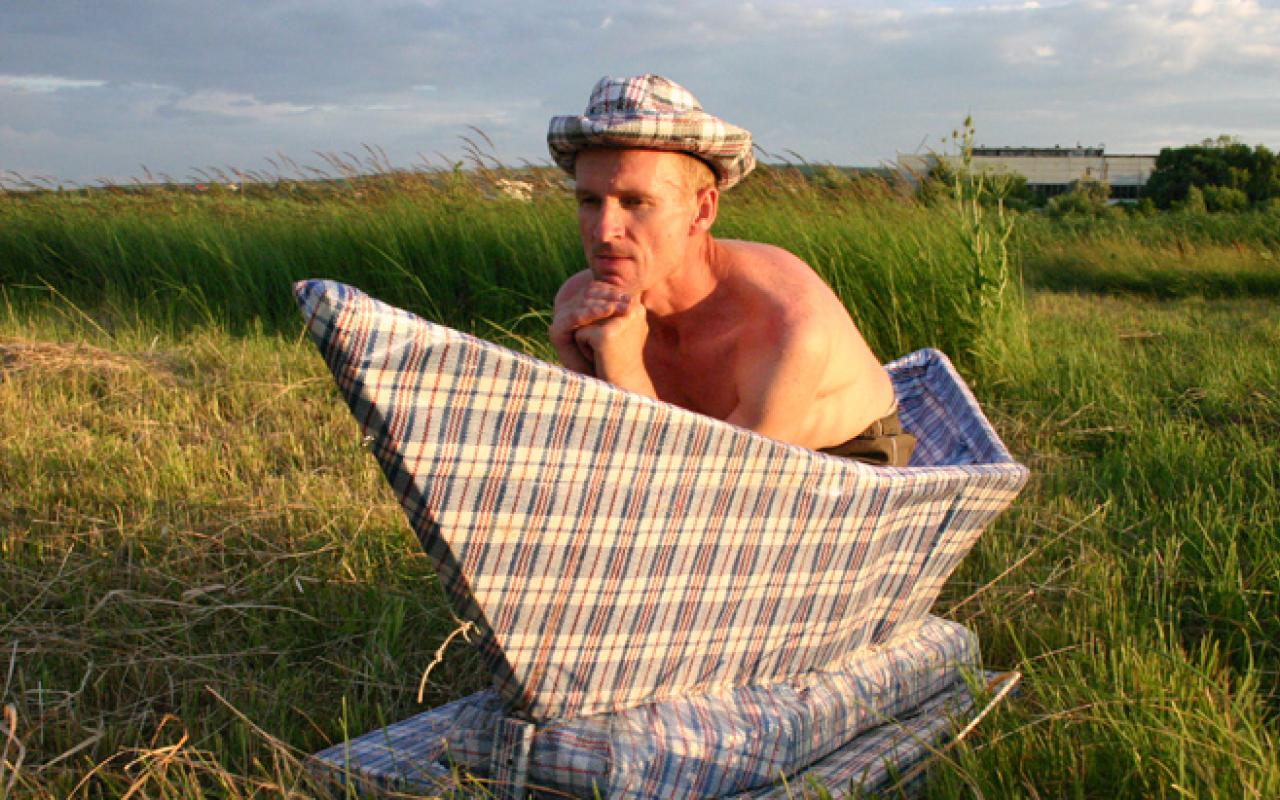 A man who is naked from the waste up sits in an oversized paper boat which stands on a green