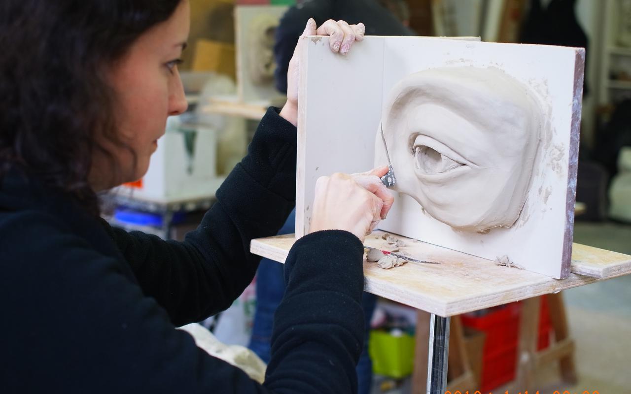 Eine Frau arbeitet an der Skulptur einer Augenpartie aus Ton. Gerade schneidet sie an der Kontur mit einem Messer ein.