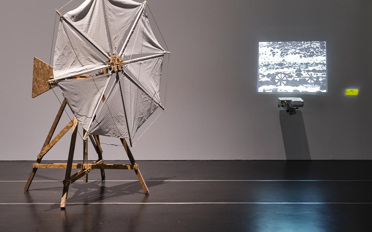 "Windmill 03 (for Walter Segal)" by James Bridle. You can see a windmill made of wood, covered with white cloth. On the right side is a small black and white projection.