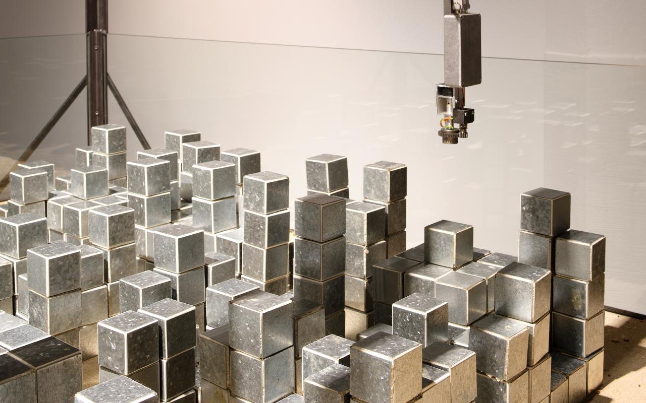 "Seek II" by Lutz Dammbeck. On view are several irregularly stacked silver cuboids. They are located in a glass box. They are moved by a device on the ceiling of the box. 