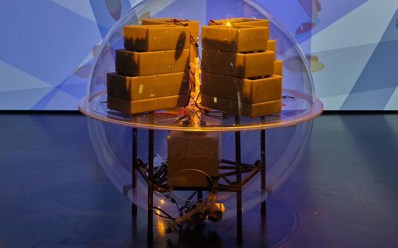 "Active Living Infrastructure: Controlled Environment (ALICE) 2.0" by Julie Freeman. A transparent sphere contains rectangular bars with a light in the center. The sphere is in front of a large screen.