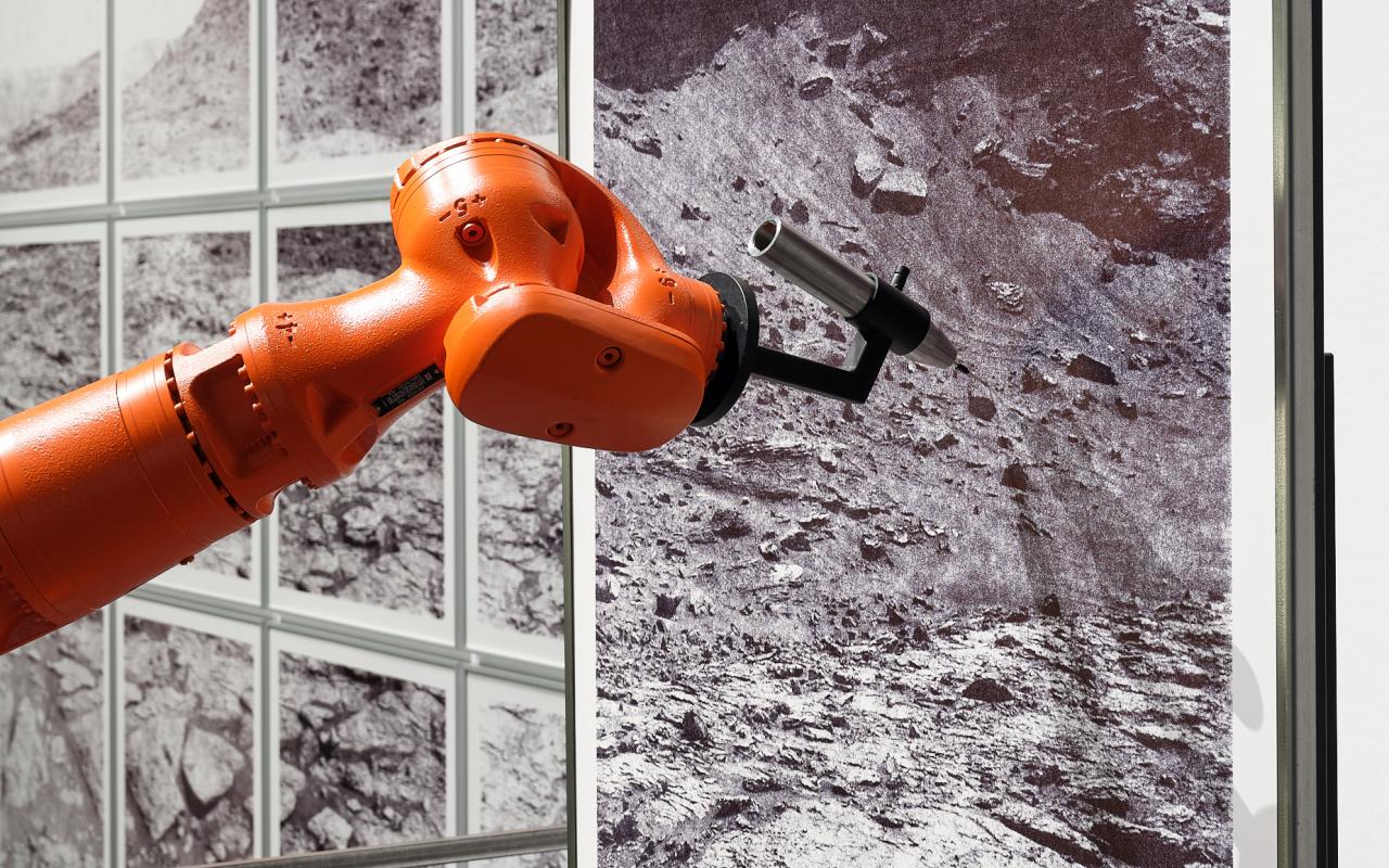 »the native picture« von robotlab. Zu sehen ist ein orangener Roboterarm, der ein schwarz-weiß Bild von einer dürren Landschaft zeichnet.