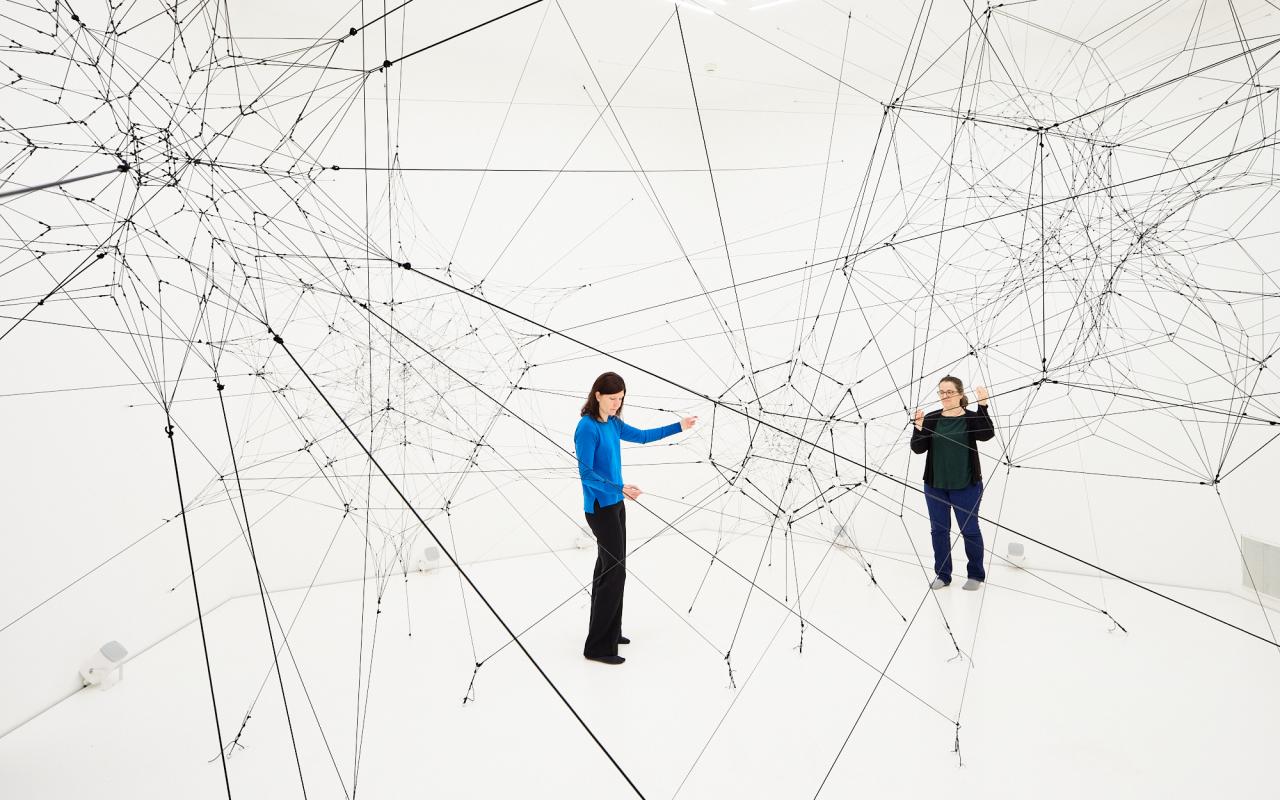 »Algo-r(h)i(y)thms« von Tomás Saraceno. Zu sehen ist ein großes Netz aus Fäden in einem weißen Raum. Zwei Personen stehen in diesem Raum und berühren diese Fäden.