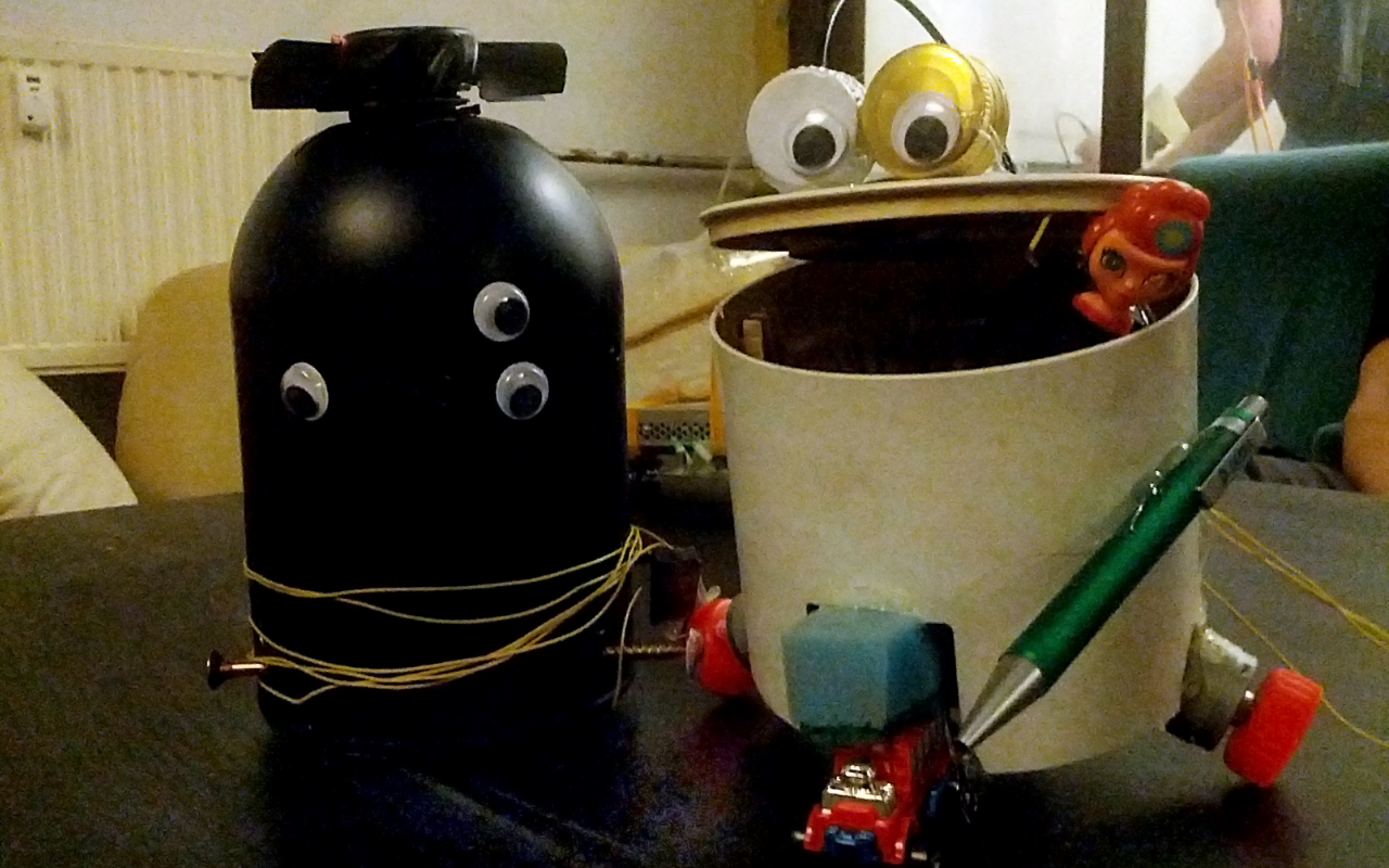 Two little robots that are made out of junk are sitting next to each other, one is black and has 3 eyes and one is made out of a bin with 2 eyes out of bottle caps, that are placed on the open mouth resp. lid of the bucket.