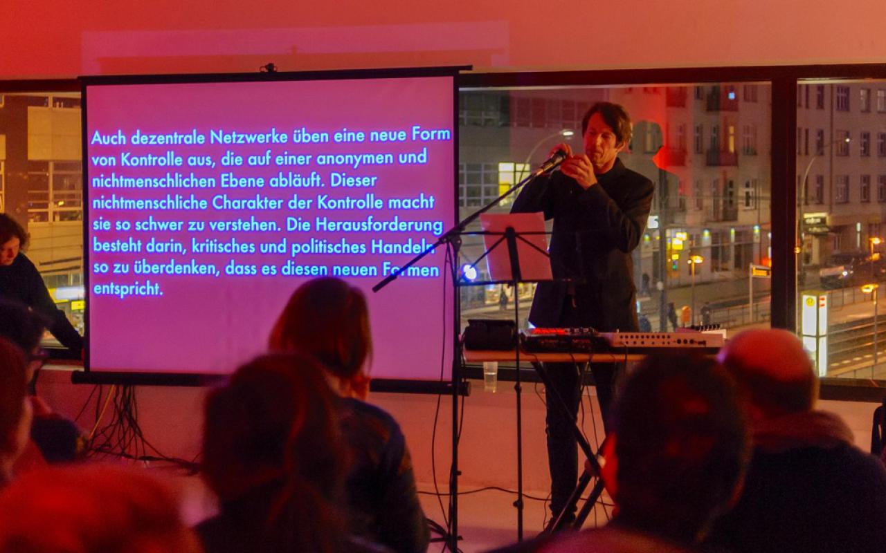 A man during his performance at the keyboards, behind him a big canvas with text being screened on it.