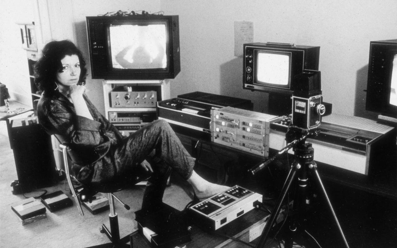 The photo is black and white and shows Ulrike Rosenbach, the media artist, in her studio in the 70s. Ulrike Rosenbach is sitting on a swivel chair and is surrounded by camera equipment and tube screens. 