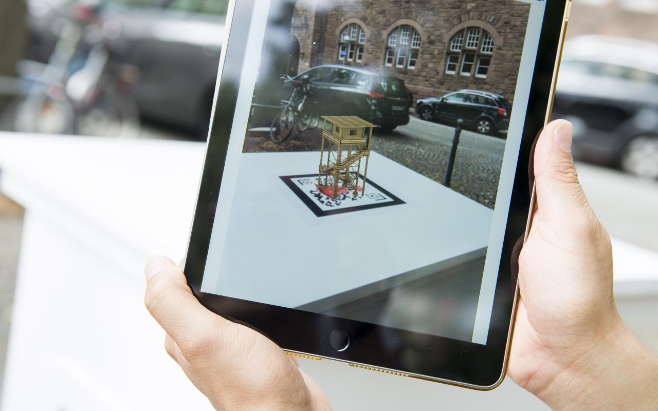 Two hands hold an iPad on which a 3D animated miniature monument can be seen over the QR code of the work »The Peoples Monuments«..