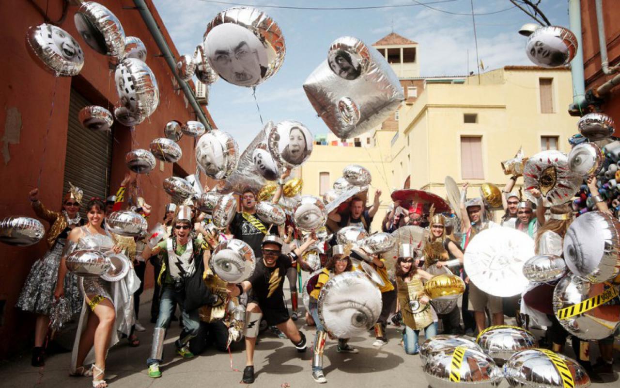 A group of men and women is standing on the street and each one is holding a silver balloon. On the balloons are pictures of faces and eyes. The people are cheering and smiling into the camera.